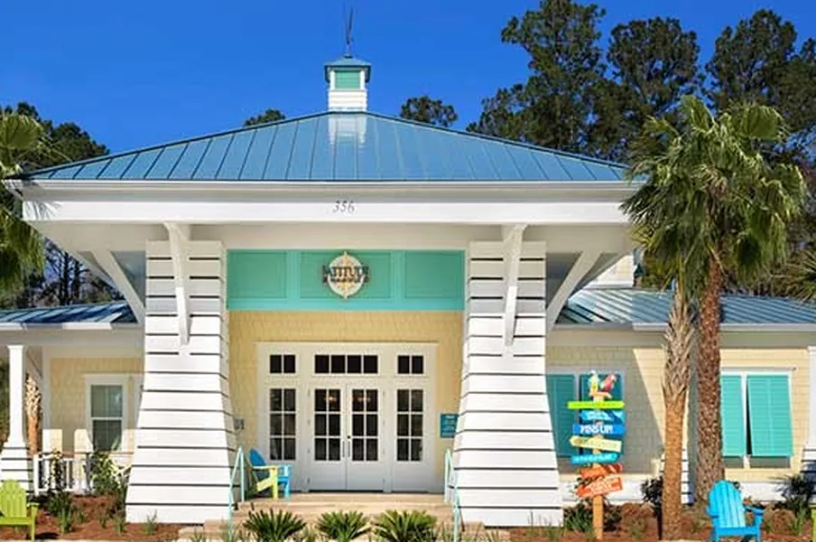 Latitude Margaritaville's Welcome Center: a blue standing-seam pyramid roof with a cupola, white lap siding on tapered posts, teal louvered shutters and a round compass-rose sign.
